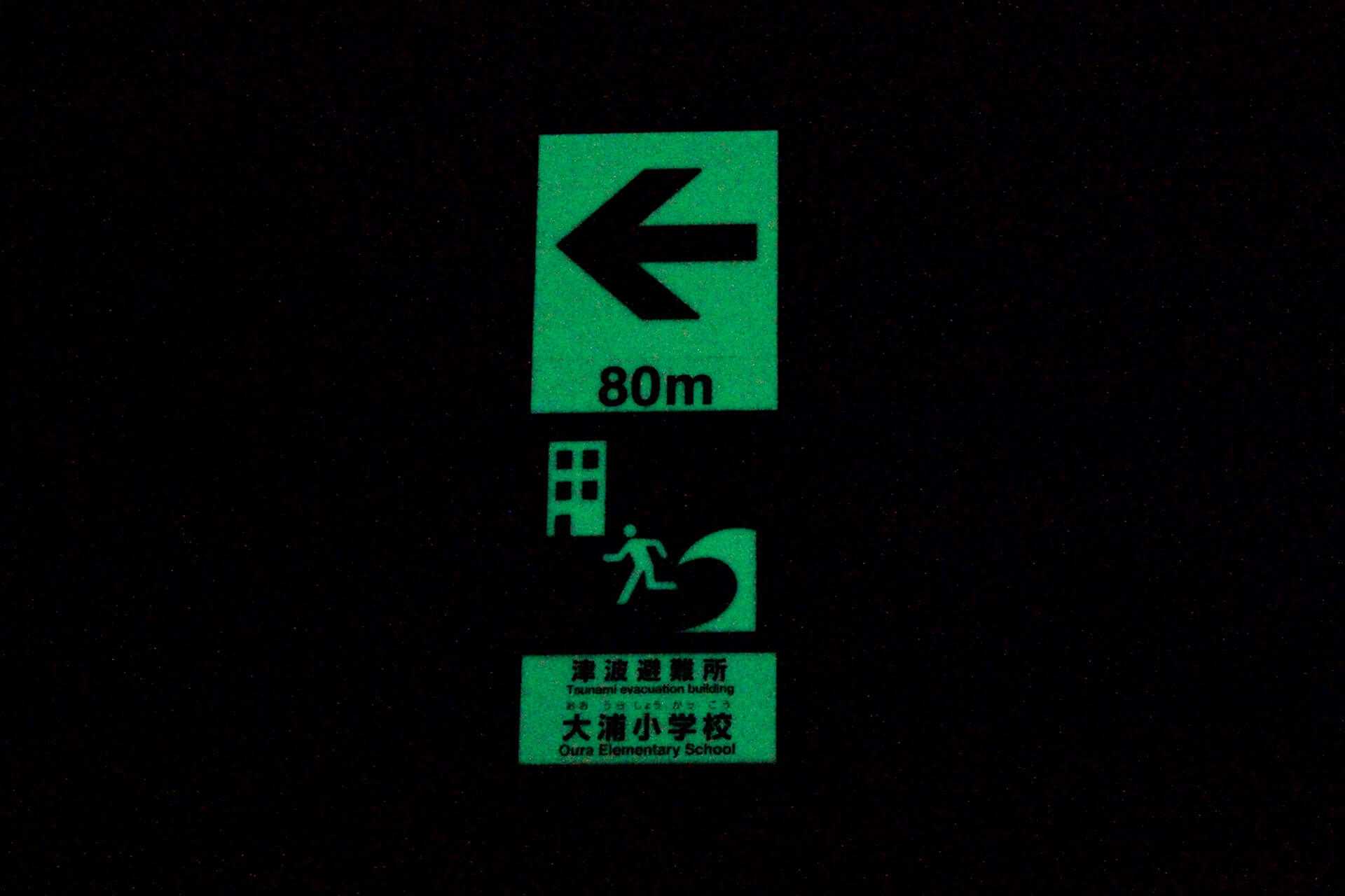 大浦小学校津波避難所へ誘導する左矢印と80メートルが標示された暗視下のハイブリッドストーン アベイラスサインプレート（高輝度蓄光式屋外用案内標示板）